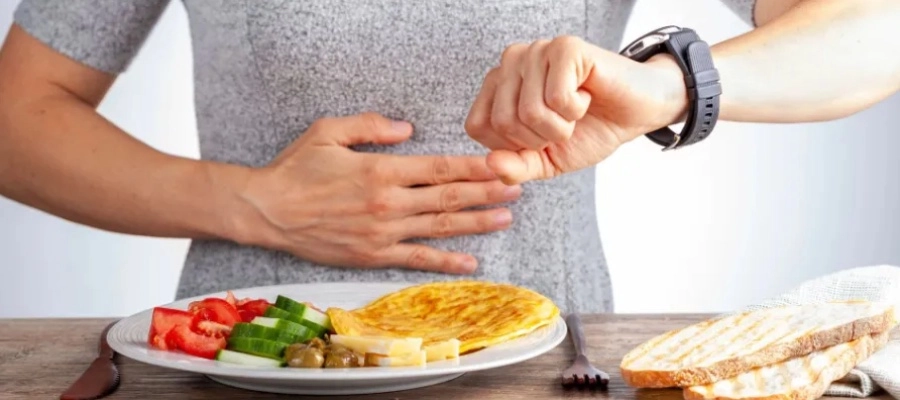 Pessoa com a mão no abdômen olhando para o relógio ao lado de um prato com ovos e legumes, representando o controle do tempo no jejum intermitente para a saúde do fígado.
