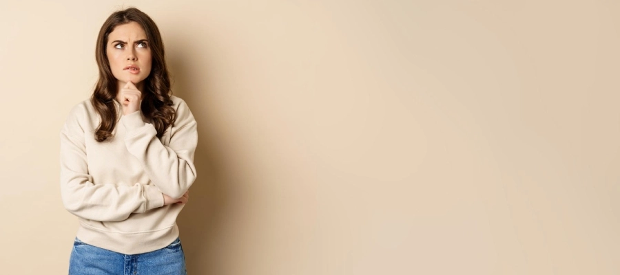 uma mulher em um suéter branco e jeans fica contra uma parede bege