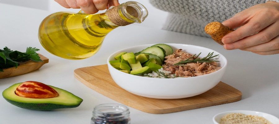Pessoa preparando uma refeição saudável, regando azeite de oliva sobre uma tigela com abacate, pepino, arroz e atum, representando uma dieta para gordura no fígado.