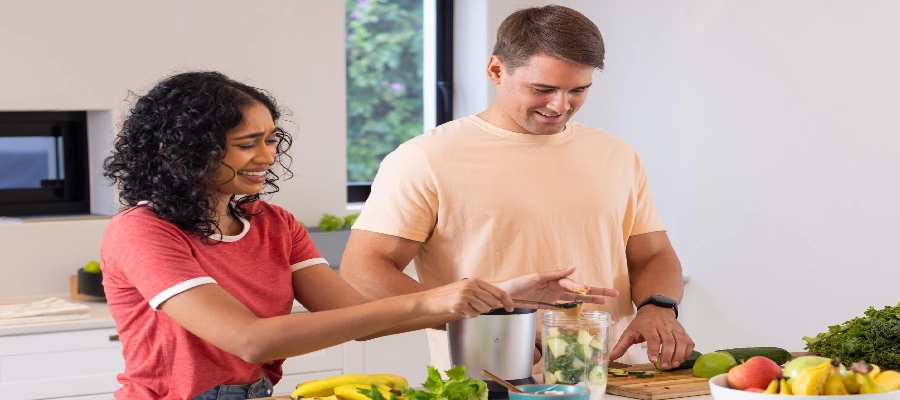 Casal sorridente preparando suco detox com frutas e vegetais frescos na cozinha, em um ambiente iluminado e saudável.