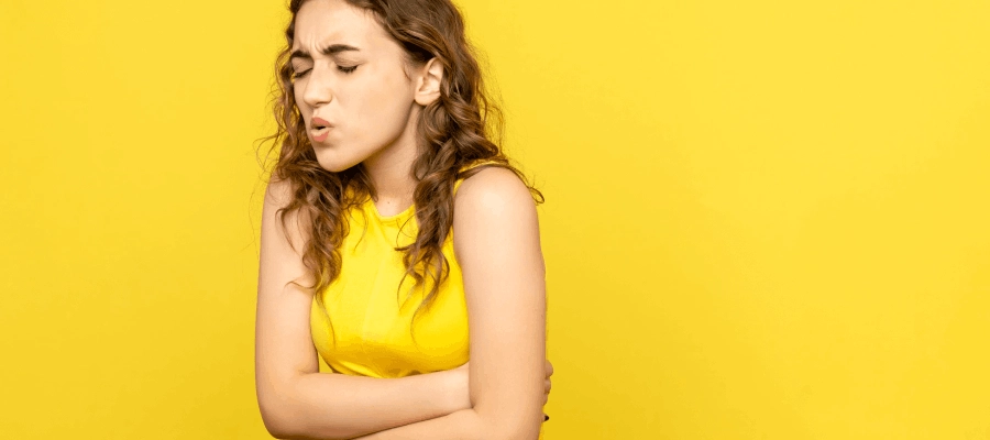 Mulher em pé em um fundo amarelo com as mãos em cima da barriga mostrando dor abdominal relacionada à inflamação no estômago e desconforto digestivo.