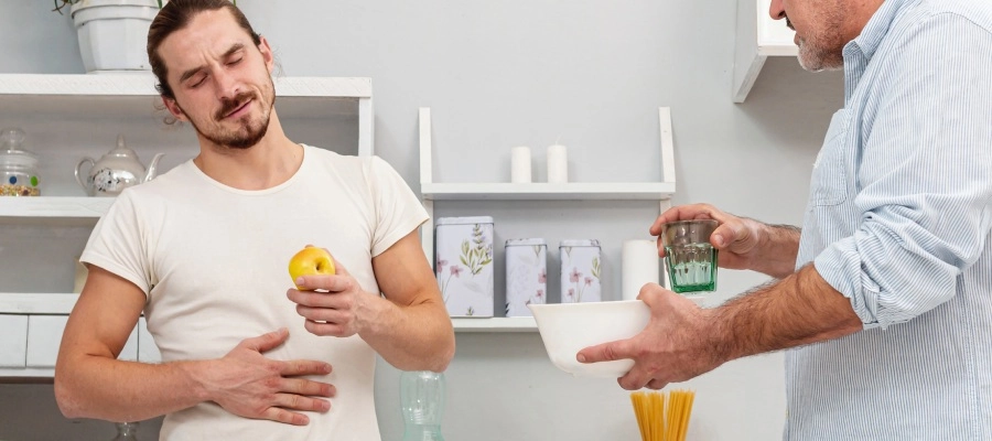Pai e filho adulto na cozinha do apartamento, com filho com dor pela acidez estomacal, comendo uma maçã, enquanto o pai oferece um copo com água.