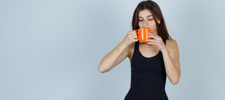 Mulher jovem com calça e camiseta pretas bebendo em uma xícara laranja, com dúvida se o termogênico faz mal para o fígado.