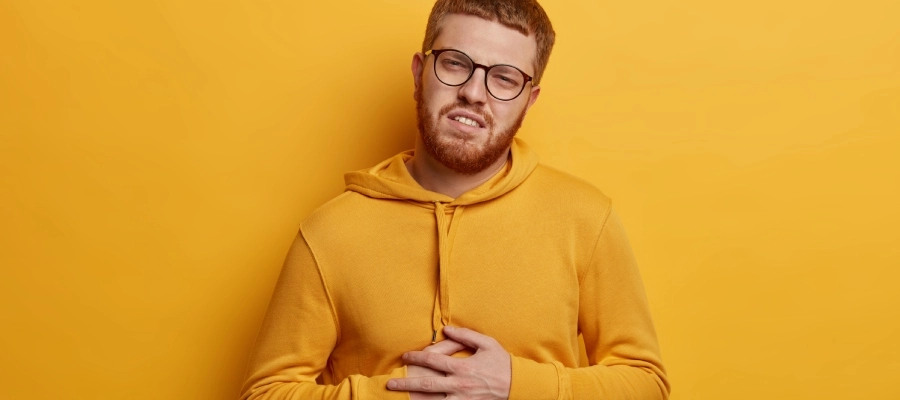 Homem ruivo de óculos e moletom amarelo segura a região do abdômen com expressão de dor e desconforto, pensando no que é bom para má digestão após uma refeição pesada.