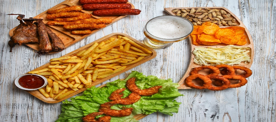 Mesa com diversos alimentos ultraprocessados, como batatas fritas, anéis de cebola empanados, linguiças, salgadinhos, pistaches e copo de cerveja.