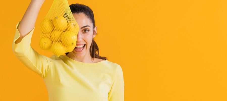 Mulher jovem com blusa amarela de manga comprida sorrindo e segurando um saco amarelo com laranjas, o que mostra uma boa alimentação para quem tem gordura no fígado.