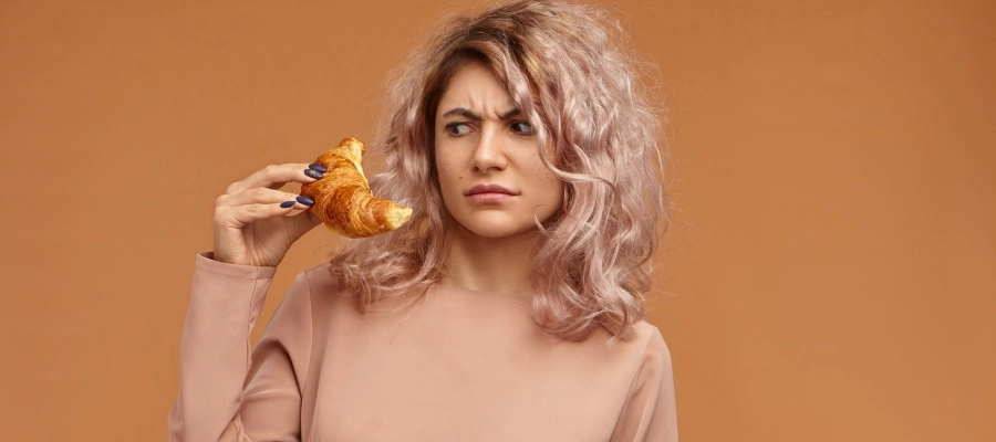 Mulher jovem de cabelo rosa segura um croissant com expressão de desconforto e frustração, ilustrando a busca por remédio para comida que fez mal após consumir um alimento que causou mal-estar.