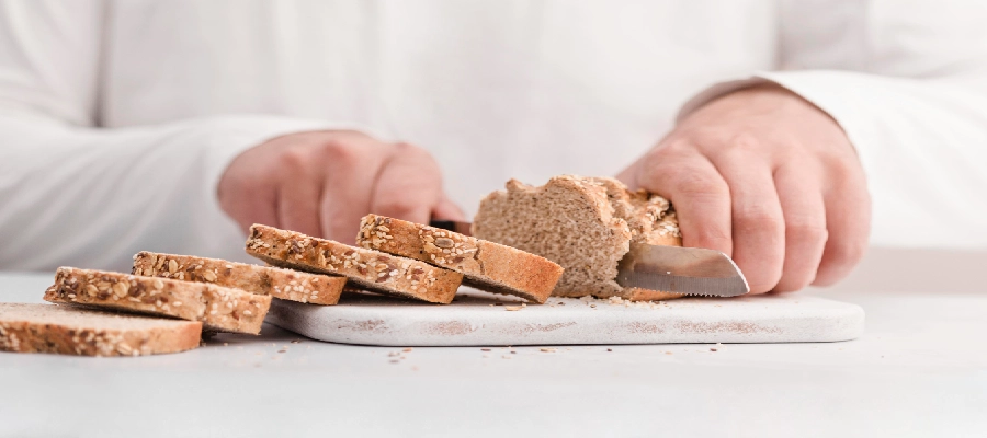 Pessoa fatiando pão integral, um dos alimentos sem glúten, em tábua branca, com fatias alinhadas em primeiro plano.