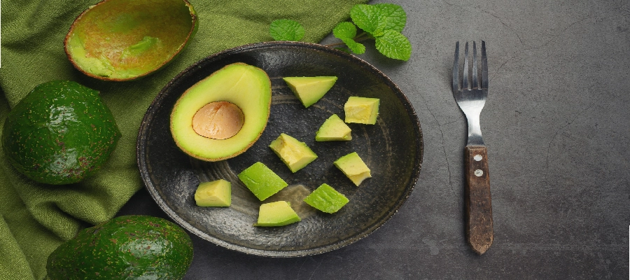 Pedaços de abacate cortados em cubos em prato preto ao lado de meio abacate com caroço. | Benefícios do abacate